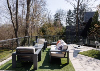 Modern outdoor patio with grey aluminum furniture, cream cushions, artificial turf, and flagstone paving, overlooking a wooded backyard.