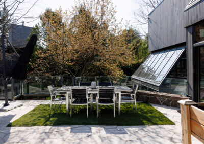 Modern outdoor dining area with a metal and wood table and chairs on artificial turf, surrounded by a flagstone patio, a closed black umbrella, and a contemporary dark-paneled house with large glass windows.