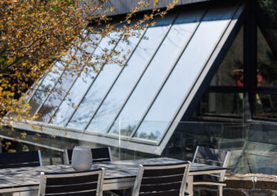 Modern outdoor dining table and chairs on artificial turf in front of a contemporary home with a large slanted glass roof and dark siding, framed by a tree with orange leaves