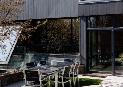 Modern house exterior with dark gray siding, large windows, and an outdoor patio featuring a dining set on artificial turf and a lounge sofa.