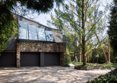 Modern house exterior with dark gray siding, stone facade, large angled windows, two-car garage, and lush natural landscaping with trees and a stone paver driveway.