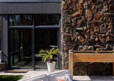 Outdoor patio with a modern cream sofa and colorful pillows against a contemporary house featuring a stone accent wall and large glass windows.