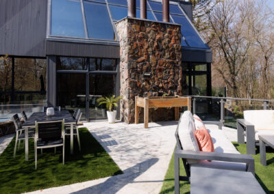 Modern home exterior with a large stone chimney, expansive glass windows, and a multi-level outdoor patio featuring dining and lounge areas with artificial turf and pavers.