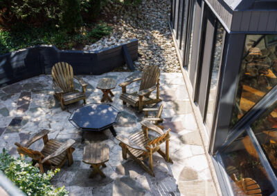 Aerial view of a flagstone patio with wooden Adirondack chairs and a hexagonal table, bordered by modern architecture and a dry river bed garden.
