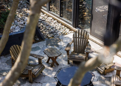 Overhead view of a stone patio with weathered wooden Adirondack chairs, a small table, and a black hexagonal fire pit, bordered by a rock garden and modern house windows.