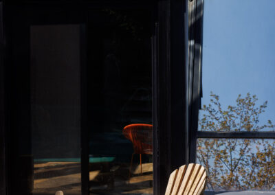 Modern dark-paneled house exterior with angular windows, a flagstone patio featuring weathered Adirondack chairs, and a fire pit, reflecting trees and sky.