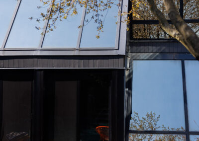 Modern house exterior with dark siding, large glass windows, and an outdoor patio featuring wooden Adirondack chairs around a fire pit, with a tree in early spring foliage.