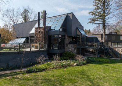Modern house with dark gray siding, extensive glass roof and skylights, stone pillars, and a large outdoor deck overlooking a green lawn with spring flowers.