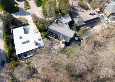 Aerial view of modern luxury homes in a treed suburban neighborhood, featuring a white-roofed contemporary house and another with glass walls and dark roof, surrounded by green landscaping.