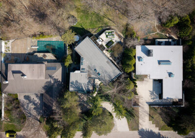 Aerial view of three distinct residential properties, including a modern flat-roof house with several outdoor patio areas, a traditional gabled-roof home, and another property with a covered swimming pool and wooden deck, all nestled within a partially wooded suburban landscape.