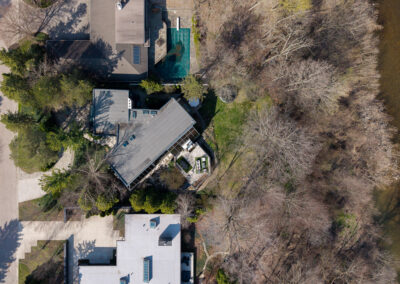 Aerial view of a contemporary riverfront home with a slanted roof, flanked by lush green lawns and bare deciduous trees, adjacent to a winding creek. Other residential properties with varied architecture and a covered swimming pool are visible in this suburban landscape.