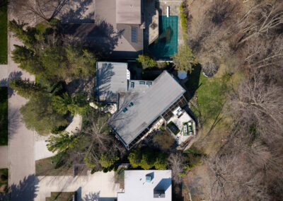 Aerial view of a modern residential property featuring a main house with a distinctive roofline, a swimming pool, and extensive landscaping with both deciduous and evergreen trees, seen from above.
