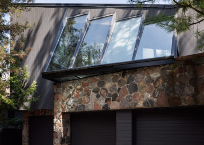 Modern house exterior with dark gray siding, natural stone facade, angled windows, and two garage doors.