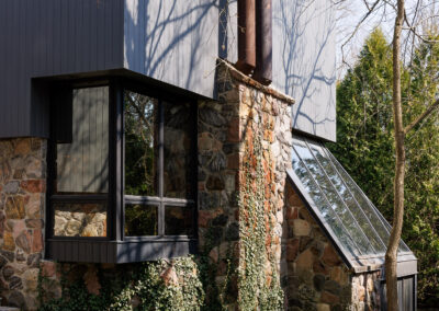 Modern house with stone and dark siding, large windows, industrial chimneys, and ivy-covered landscape under a clear sky.