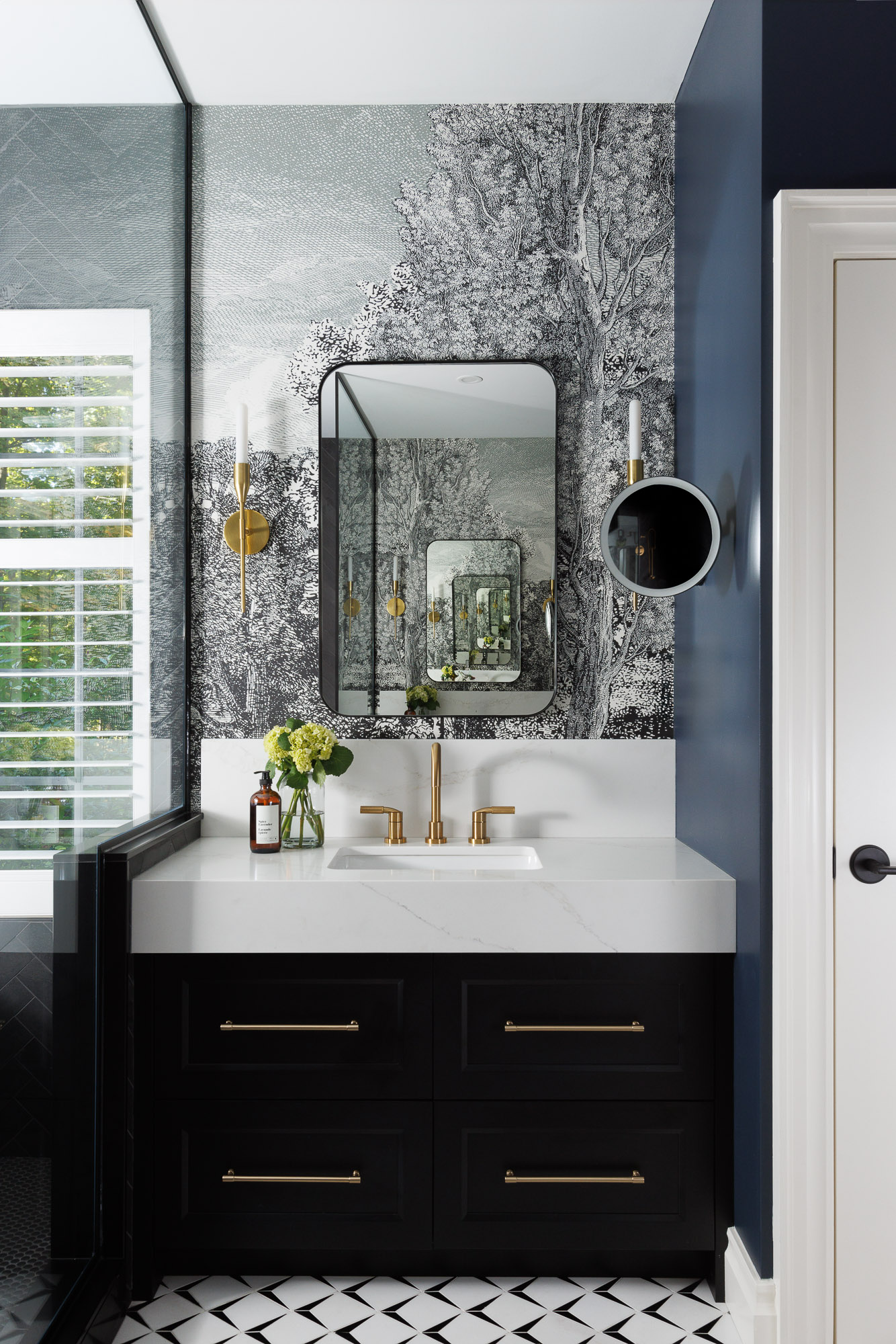 black-vanity-woodland-wallpaper-modern-residence Modern bathroom vanity featuring black cabinetry with gold hardware, marble countertops, and detailed woodland-themed wallpaper under brass wall sconces.