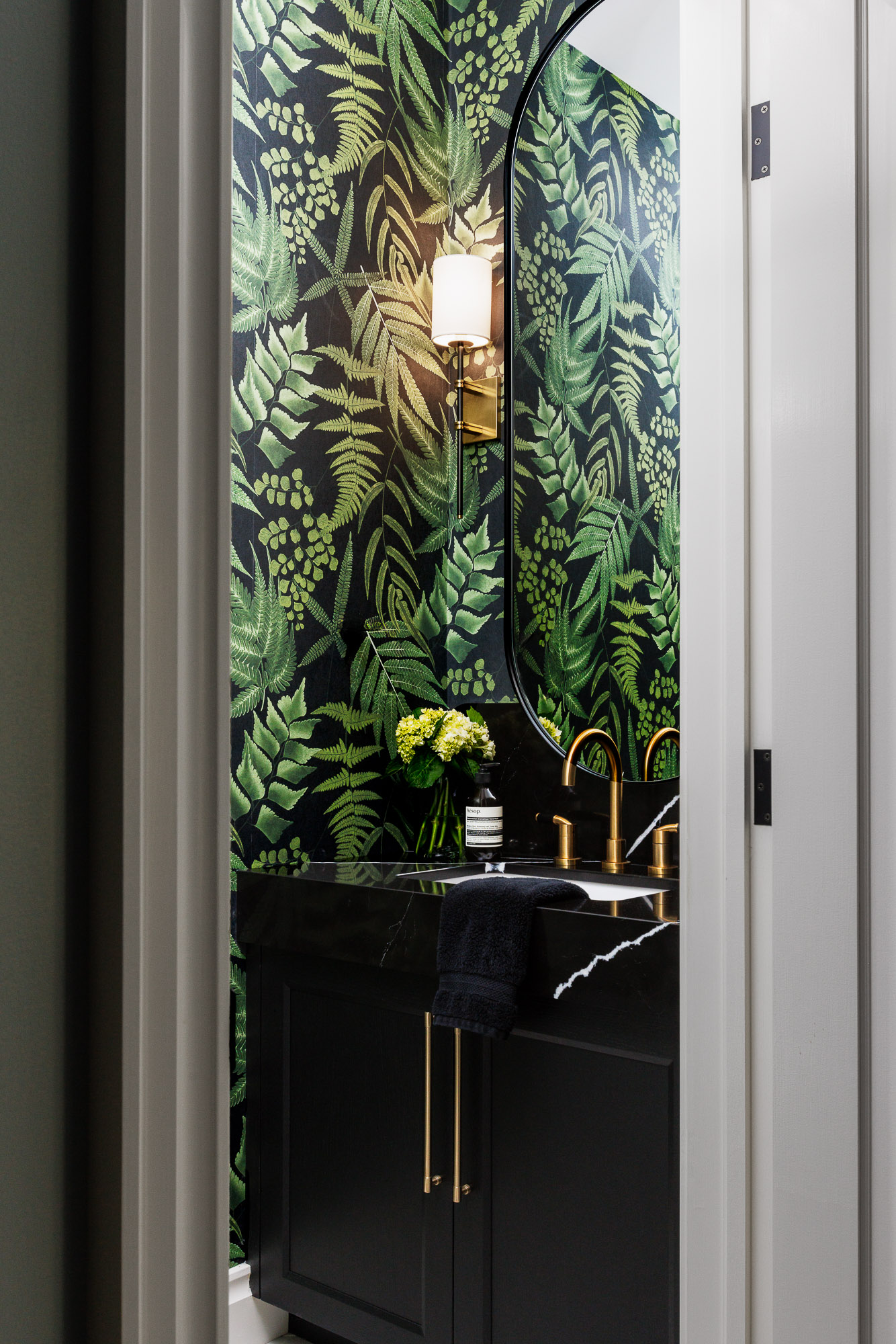 Luxury Powder Room with Botanical Wallpaper and Brass Accents Professional architectural photograph of a moody powder room featuring green fern-patterned wallpaper, a black marble vanity with long brass handles, and a gold-toned faucet.