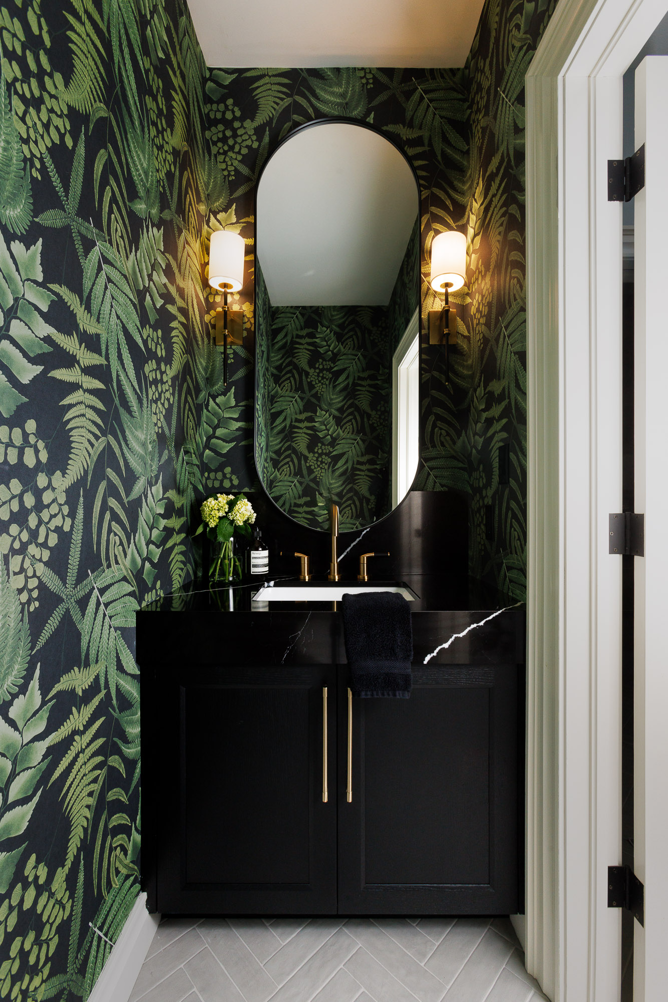 Modern Powder Room Interior with Herringbone Flooring Straight-on architectural view of a powder room with fern wallpaper, a black vanity, twin gold sconces, and a light gray herringbone tile floor.