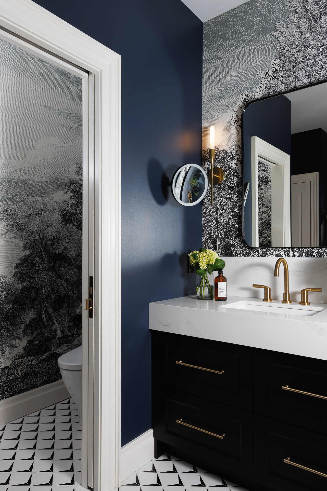 navy-blue-powder-room-suburban-estate-monochrome-wallpaper Suburban estate powder room with monochrome landscape wallpaper, navy blue accent wall, brushed gold hardware, and geometric tile flooring.