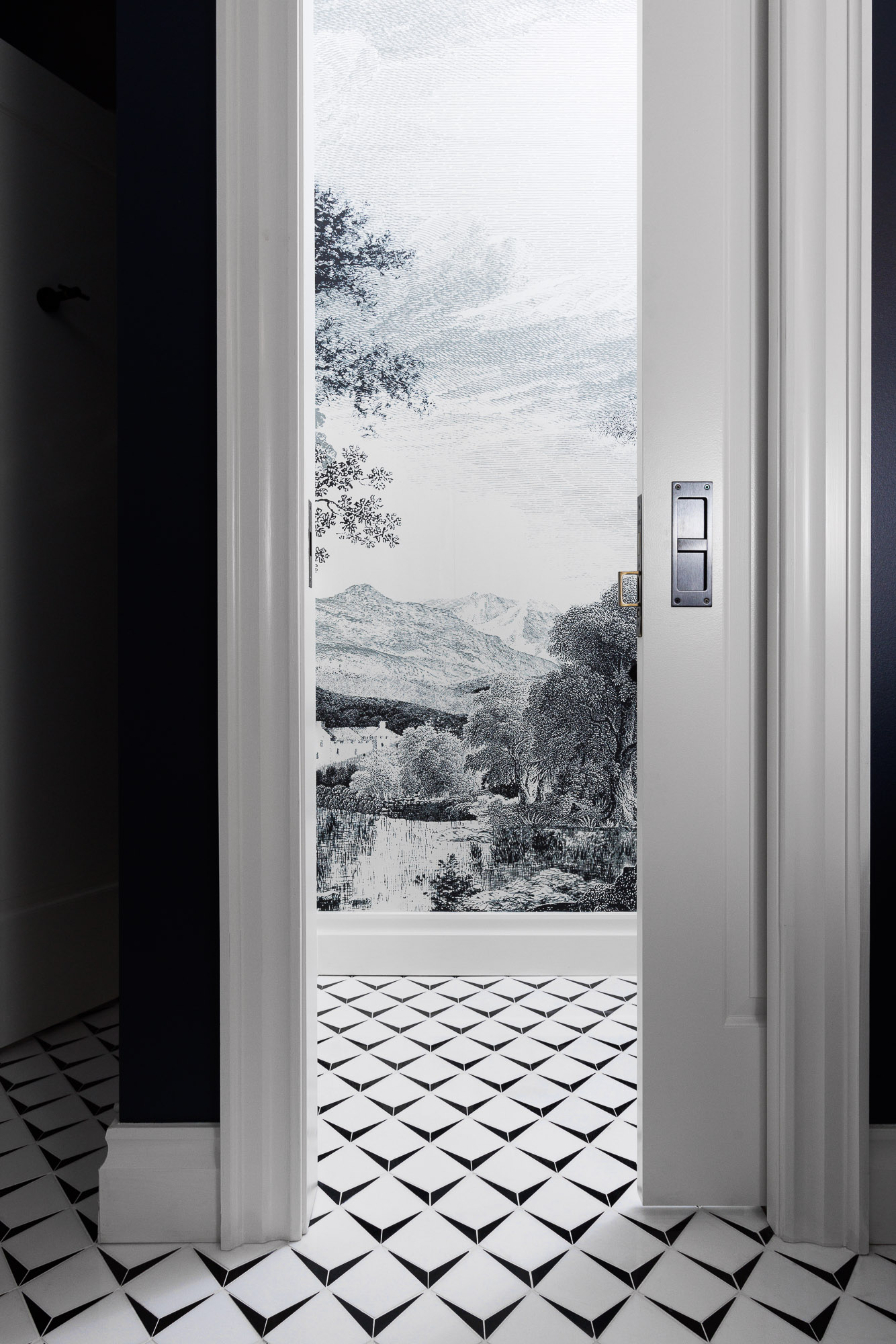 monochrome-powder-room-pocket-door-suburban-estate A white sliding pocket door opens to a powder room featuring monochrome geometric floor tiles and a scenic landscape wall mural.