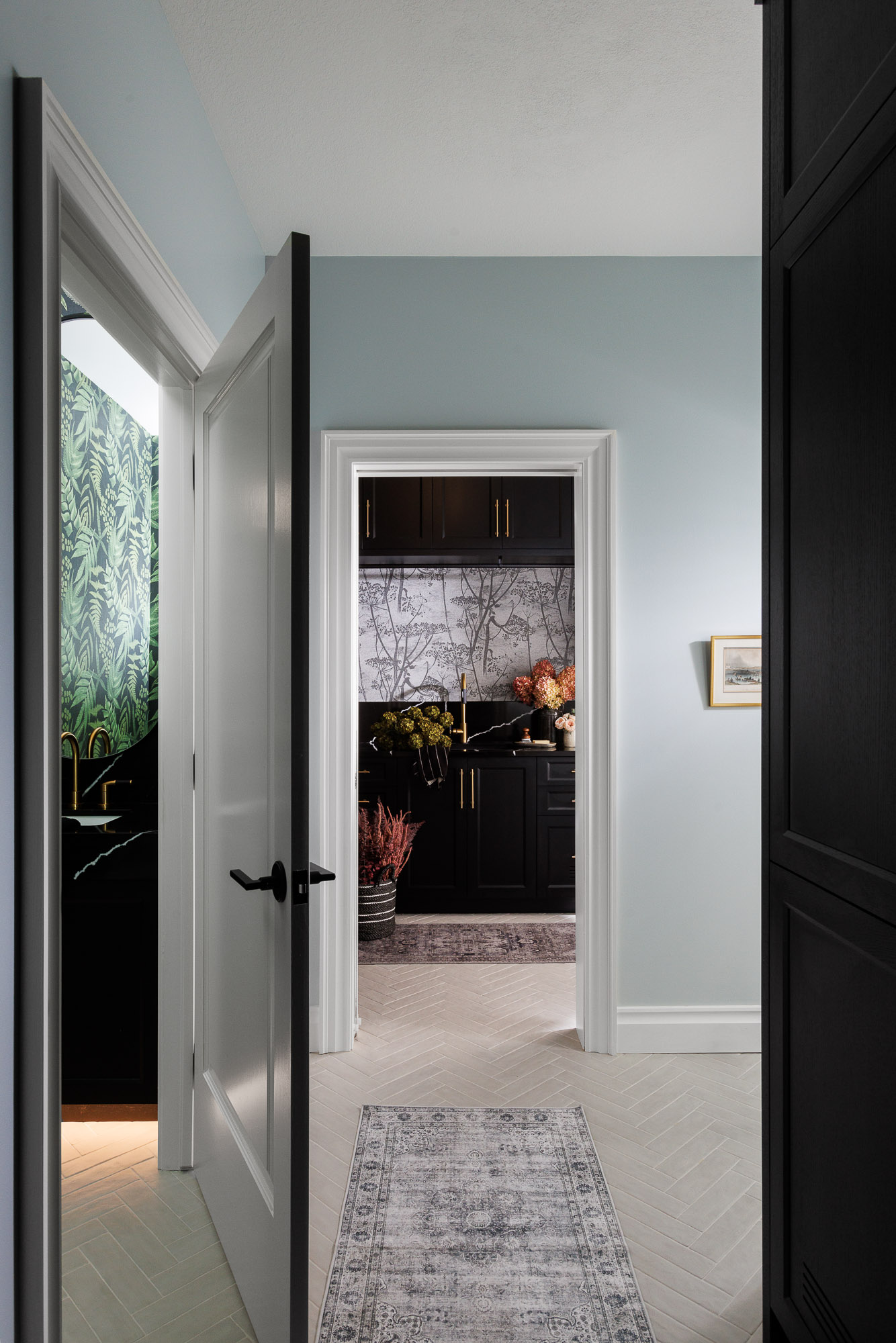 Laundry Room Entry with Botanical Wallpaper A view through a white door frame into a laundry room featuring black cabinetry, floral wallpaper, and a light-colored herringbone floor.