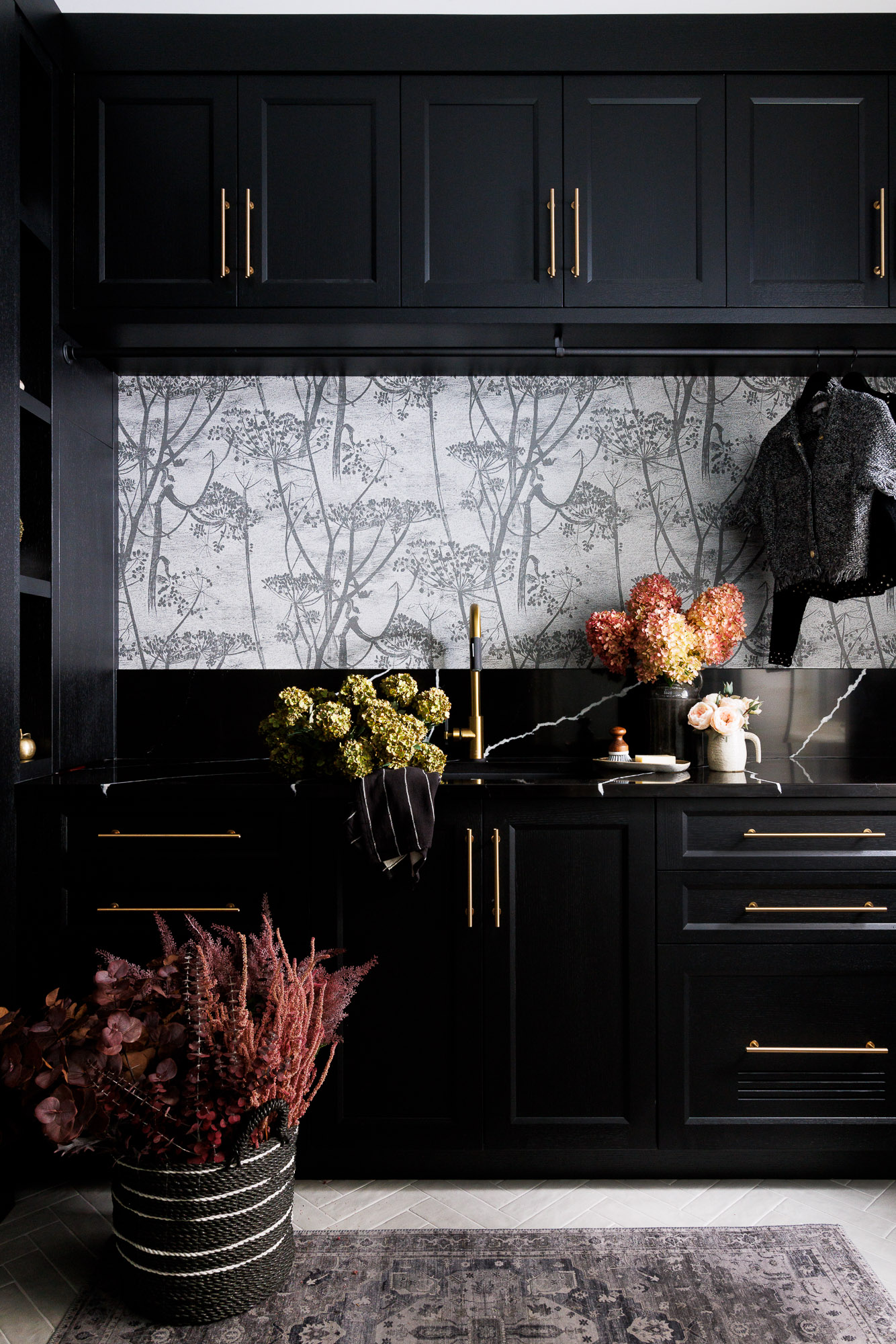 Black Laundry Cabinetry and Floral Mural A detailed shot of black laundry room cabinets with a gold faucet and black marble-look backsplash, set against a botanical mural.