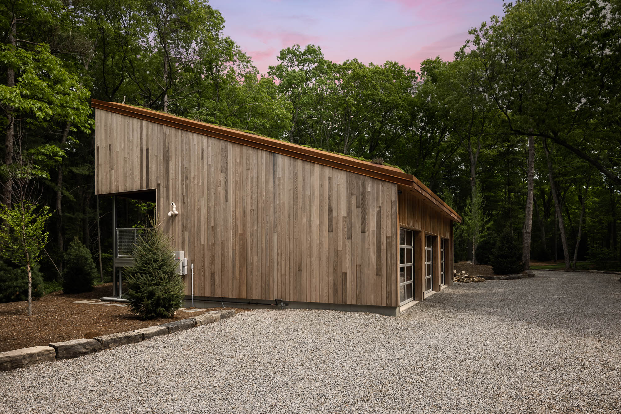 Modern Wood Building and Gravel Driveway View of a contemporary wood-clad garage and gravel driveway leading into a forested property.