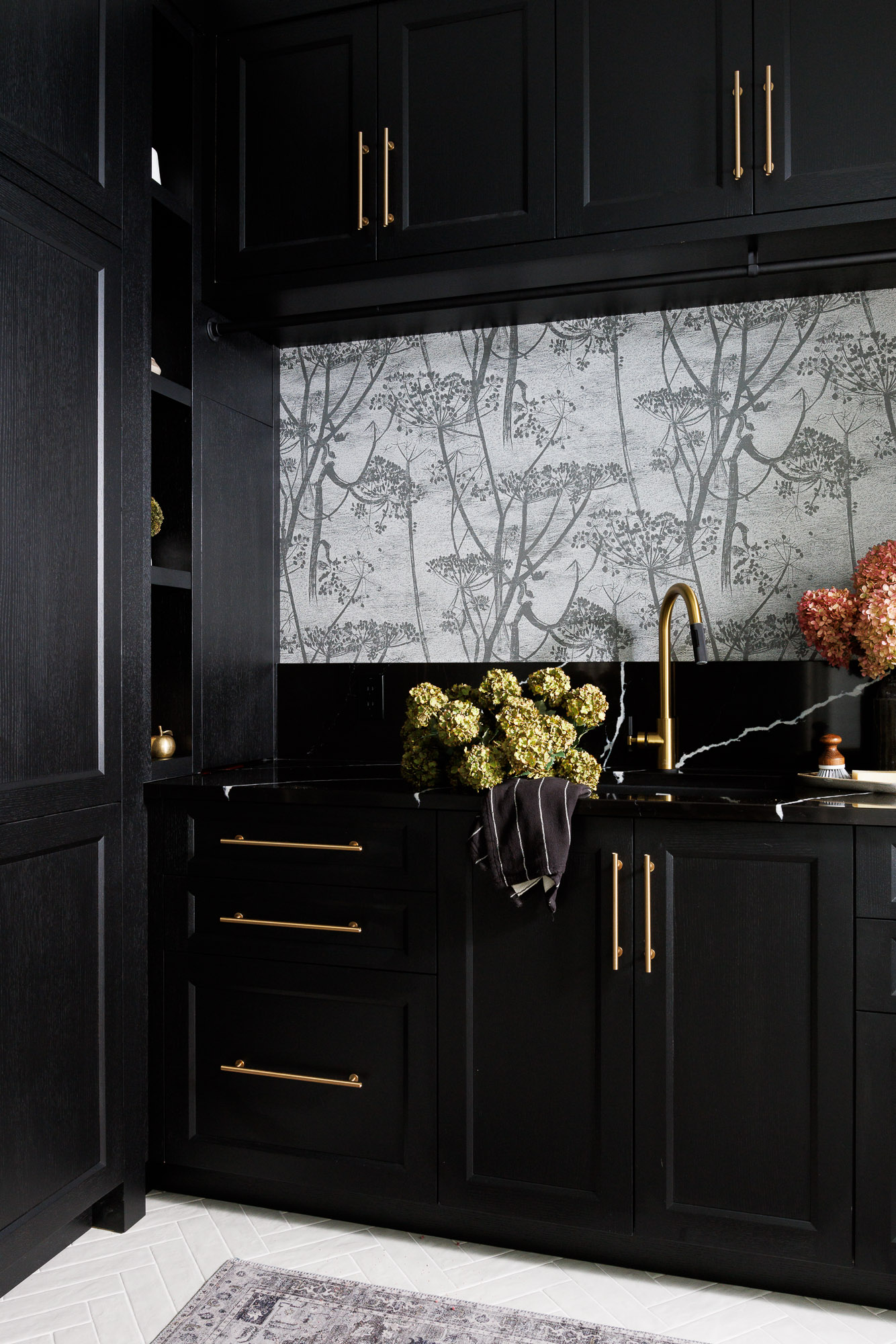 Gold Faucet Detail in Botanical Laundry Room Close-up of a brushed gold laundry room faucet and black stone countertop with a herbaceous wallpaper background and a bouquet of hydrangeas.