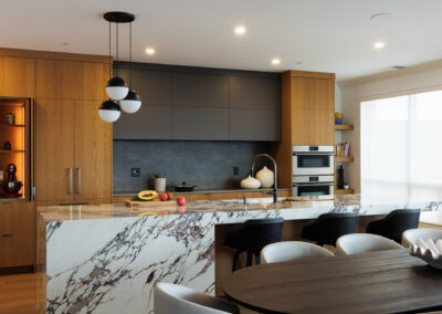 Modern kitchen with large white and brown marble waterfall island, light wood cabinetry, dark grey upper cabinets, and black spherical pendant lights over the island.