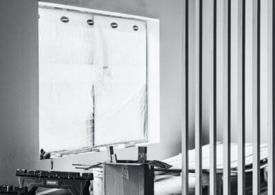 Monochrome image of an interior construction site with a plastic-covered window, modern wooden slat room divider, and Stanley saw horses on a concrete floor.