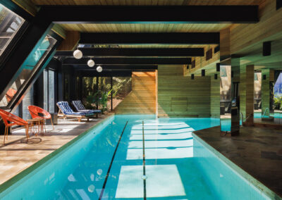 Modern indoor swimming pool with wooden walls, large glass windows, lounge chairs, and natural light
