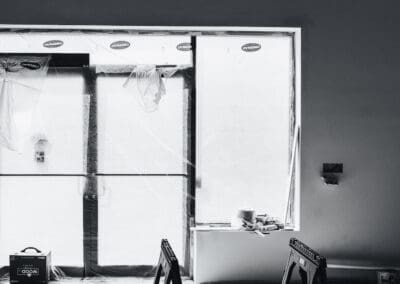 Black and white photo of an indoor construction site with two black saw horses on a concrete floor in the foreground, facing large windows covered with 'DYNAMIC' branded protective plastic sheeting. A 'WOOD design' box is visible on the left.
