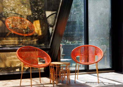 Two vibrant orange Acapulco-style chairs and a matching side table positioned beside an indoor swimming pool and large glass windows.