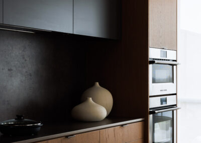 Modern kitchen with dark gray upper cabinets, natural wood lower cabinets, black countertop, and integrated double ovens.