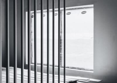 Black and white interior of a construction site with vertical wooden slats, a large window covered in plastic, and building materials on a concrete floor.