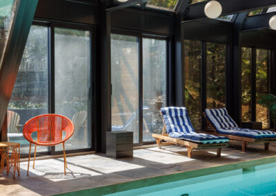 Modern indoor swimming pool with lounge chairs, an orange accent chair, and large glass windows offering a garden view.