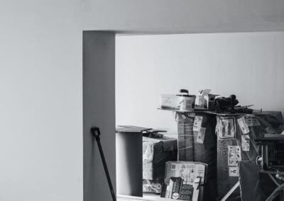 Black and white photo of an unfinished room or construction site with stacked boxes of building materials, tools, and a shovel leaning against a freshly painted wall.