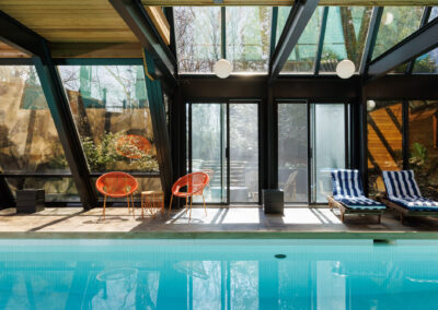 Modern indoor swimming pool with glass roof, expansive windows, and a lounge area featuring orange chairs and striped recliners, bathed in natural light.