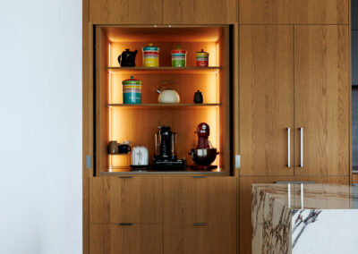 Modern kitchen with built-in light oak wood cabinetry, an illuminated appliance pantry featuring a coffee machine, toaster, and stand mixer, alongside a white marble kitchen island.