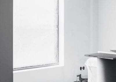 Monochrome image of an interior room under construction, featuring a large window covered with 'DYNAMIC' brand protective plastic film, unfinished white walls, exposed electrical outlets, and stacks of trim boards on the floor.