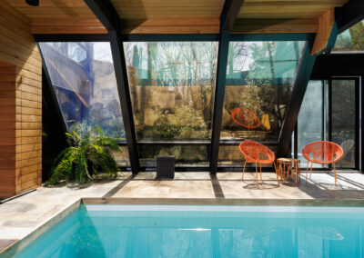 Modern indoor-outdoor swimming pool with glass enclosure, wood paneling, flagstone patio, and two vibrant orange chairs