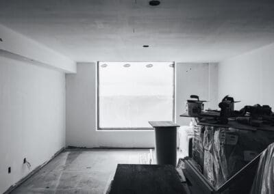 Black and white photo of an unfinished room during construction, featuring white walls, a large window covered in protective plastic, and stacked building materials.