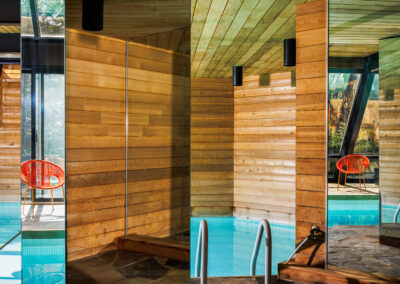 Indoor swimming pool with modern wooden walls, flagstone floor, and mirror reflections, showing a vivid orange chair through a window.