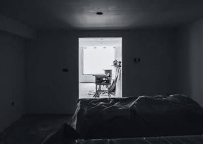 Black and white view of a dark, unfinished room with construction tools visible through a bright doorway and a plastic-covered object in the foreground.