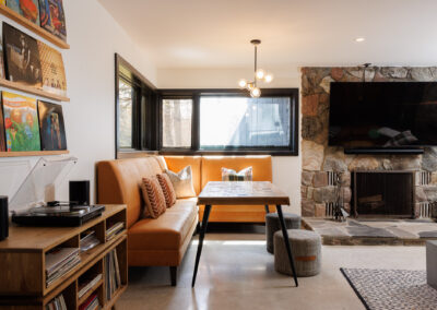 A modern living room featuring a warm tan leather L-shaped banquette, a wooden dining table, a stone fireplace with a mounted TV, a record player, and shelves displaying vinyl records.