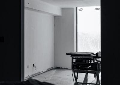 Black and white interior view of a room under construction, showing bare walls, exposed electrical outlets, a concrete floor, and a table saw positioned near a large window covered with 'DYNAMIC' branded protective film.