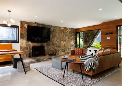 Rustic modern living room featuring a stone fireplace with a mounted TV, a brown leather sofa, geometric rug, and a game table with an orange bench.