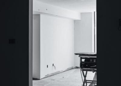 Monochrome image of an unfinished room under construction, viewed through a dark doorway, featuring exposed wiring and a table saw.