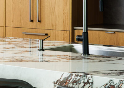 Modern kitchen island with light wood cabinets, white marble countertop, dark gooseneck faucet, and bar stools.