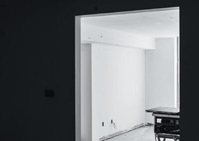 Black and white image of an unfinished room with white walls and exposed electrical outlets, viewed through a dark doorway, suggesting construction or renovation. A workbench is partially visible on the right.