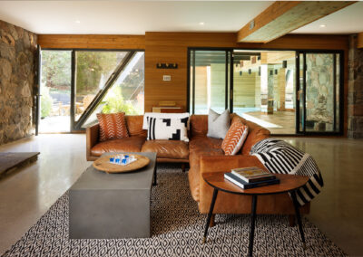 A stylish modern living room featuring a large tan leather L-shaped sectional sofa, geometric patterned rug, stone accent walls, wood paneling, and large windows connecting to outdoor spaces.